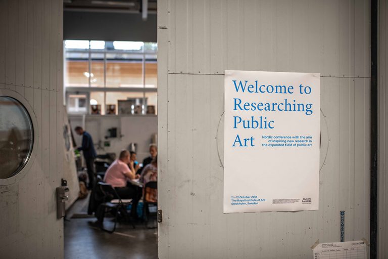 Halvöppen dörr där man ser folk sitta och prata i grupper och skriva på block. På en stängd dörr bredvid står det 