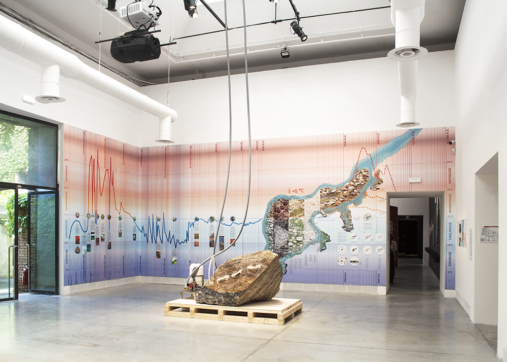 En stor sten med slangar i ligger mitt i ett rum. Bakom finns grafer och tidslinjer i olika färger.