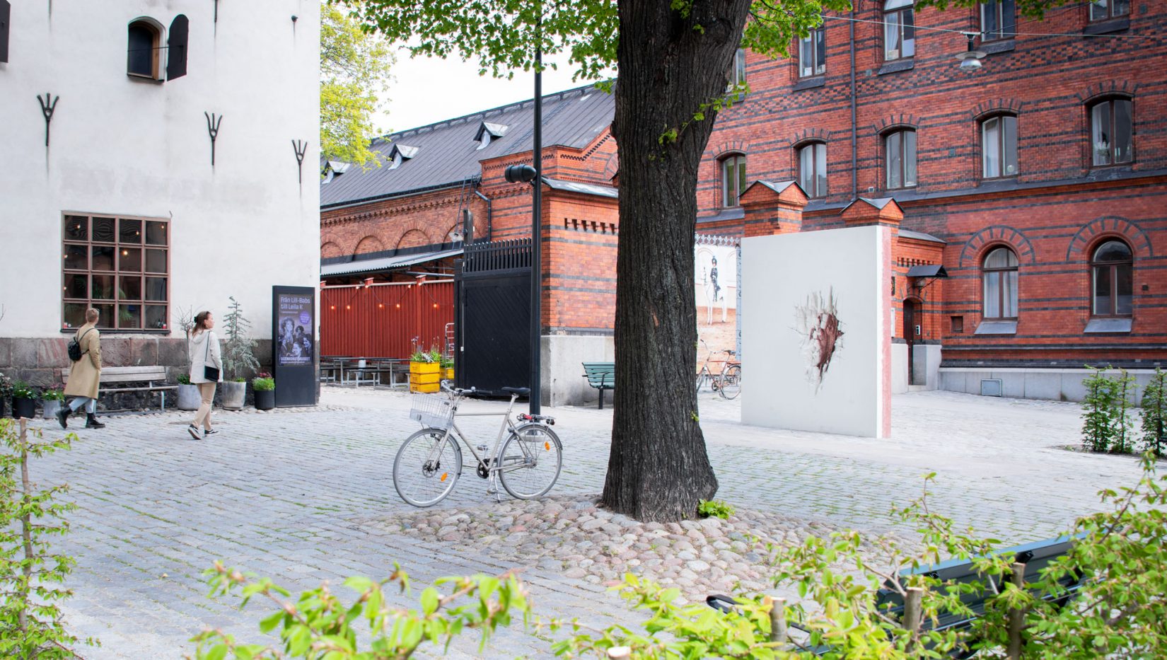 Öppet torg med ett träd i mitten. Människor går förbi och tittar på verket Brottyta (vit/röd) där det sticker upp ur marken.